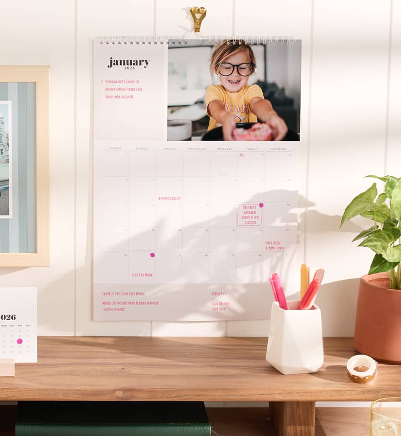  Custom Wall Calendar hung over a wooden desk with plants and a cup filled with pens. Click to shop custom wall calendars.