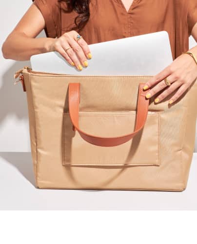 woman putting a laptop inside of a tan tote bag featuring light brown straps.