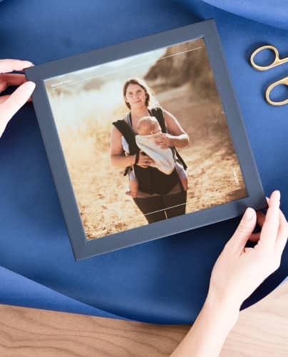 black-framed photo of woman hiking while holding baby. Framed photo is on top of blue wrapping paper with ribbon and scissors nearby.