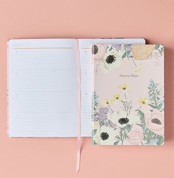 open a5 Softbound Planner showing weekly layout. One closed A5 Softbound planner closed on top featuring floral design. Click to shop Dated A5 Softbound Planners.